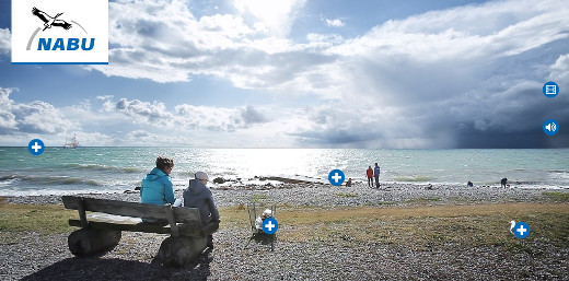Frau und Kind sitzen auf einer Bank am Strand, daneben steht ein Müllkorb, weiter entfernt eine Möwe und weitere Menschen, am Horizont fährt ein Kutter.