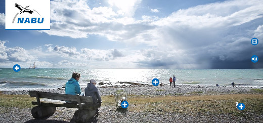 Frau und Kind sitzen auf einer Bank am Strand, daneben steht ein Müllkorb, weiter entfernt eine Möwe und weitere Menschen, am Horizont fährt ein Kutter.
